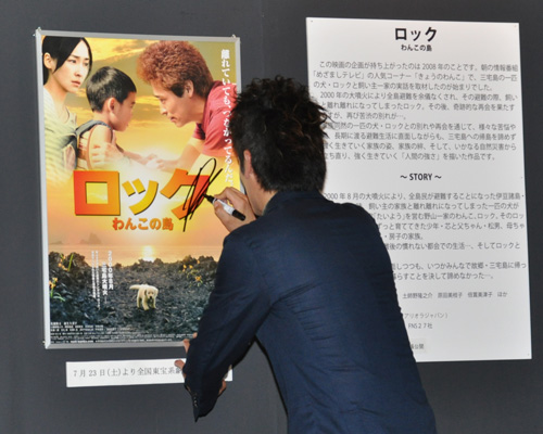 「三宅島パネル展」に展示されている『ロック 〜わんこの島〜』のポスターにサインをする佐藤隆太